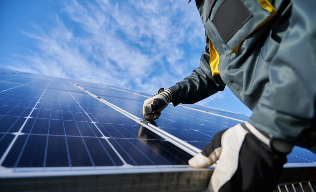Technicien ganté fixant des panneaux solaires photovoltaïques bleus sous un ciel clair. Installation d'énergie renouvelable.