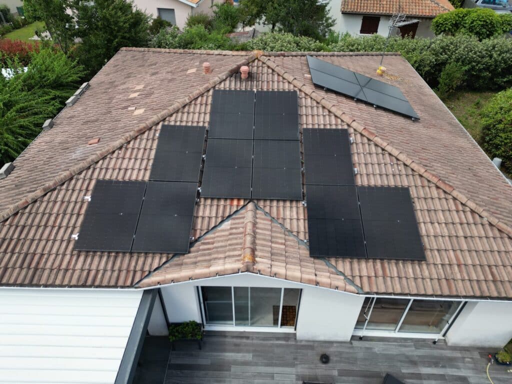 Toit de maison en tuiles terre cuite avec nombreux panneaux solaires noirs installés. Entouré de verdure, une terrasse en bois est visible.