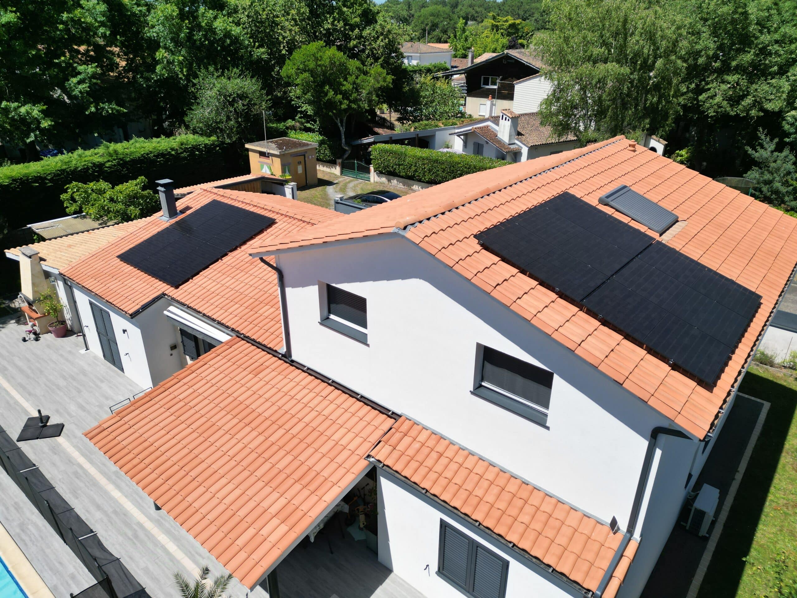 Maison moderne vue aérienne : toit de tuiles orange, 2 systèmes de panneaux solaires noirs. Murs blancs, terrasse, verdure et piscine.