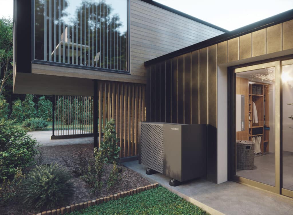 Pompe à chaleur Atlantic grise devant une maison moderne en bois et noir. Jardin verdoyant. Intérieurs visibles: bureau à l'étage, buanderie avec unité Atlantic.