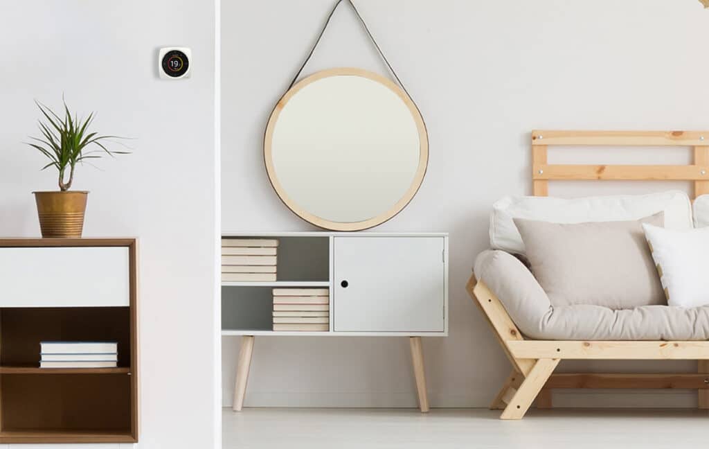 Thermostat intelligent 19° sur mur blanc, plante, meuble bois-blanc, miroir rond, canapé futon en bois clair et coussins moelleux.
