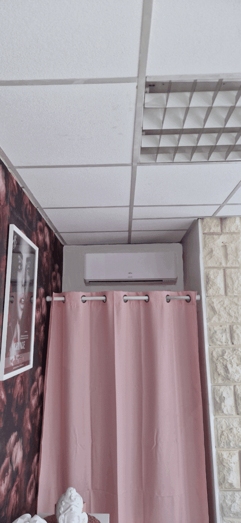 Intérieur avec plafond à dalles, climatiseur Fujitsu, rideau rose à œillets devant une niche, affiche « GRUNGE » et mur en pierre blanche.