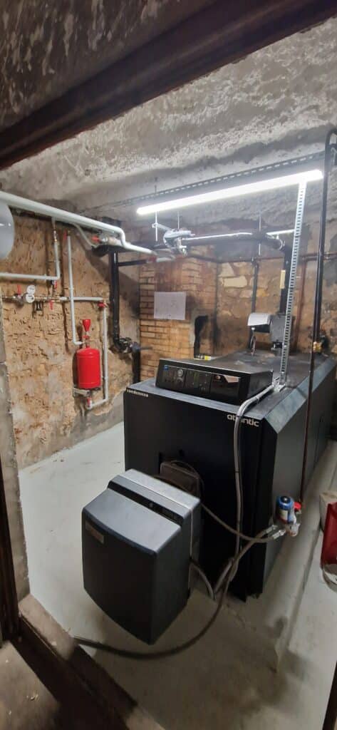 Chaudière biomasse Atlantic Condenseo noire installée dans une chaufferie aux murs de pierre et plafond isolé, avec tuyauterie blanche et réservoir rouge.